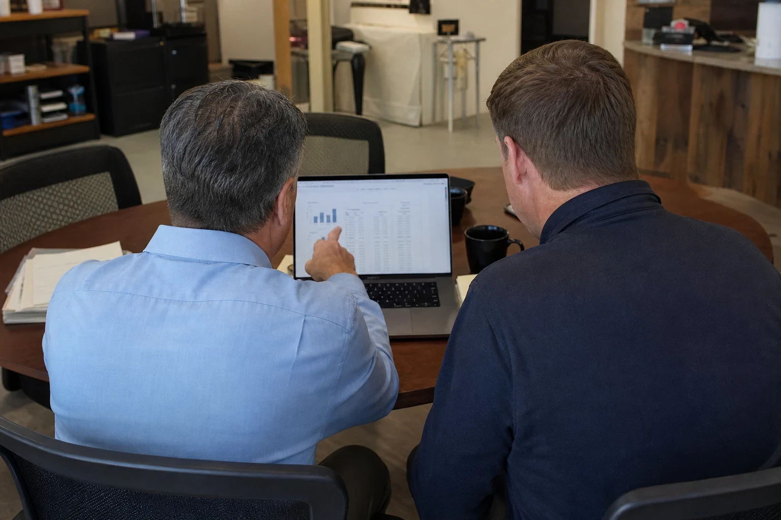Two men seen from behind at a round conference table in a storefront office, one in a light blue shirt pointing at a chart on a laptop screen