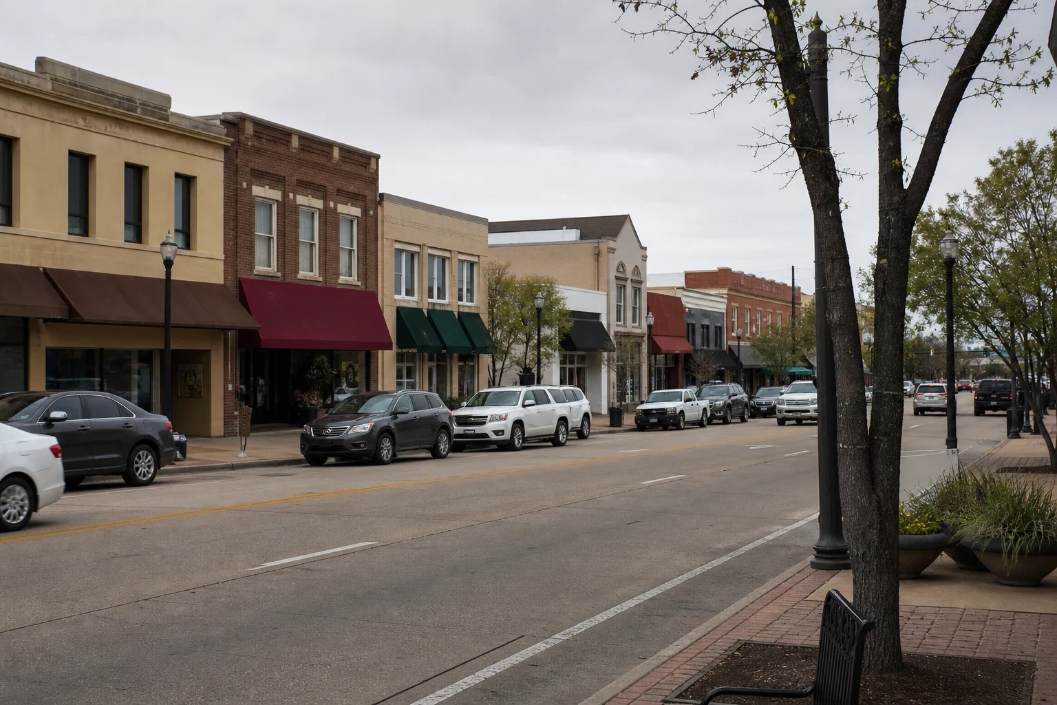 Overcast main street with brick and stone storefronts under colored awnings, cars parked along the curb, street lamps and a brick sidewalk with trees