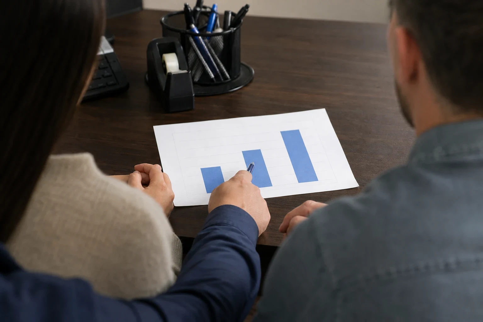 Over-the-shoulder view of a couple at a dark wood desk as an agent points a pen at a printed bar chart, with a tape dispenser, pen cup and keyboard nearby